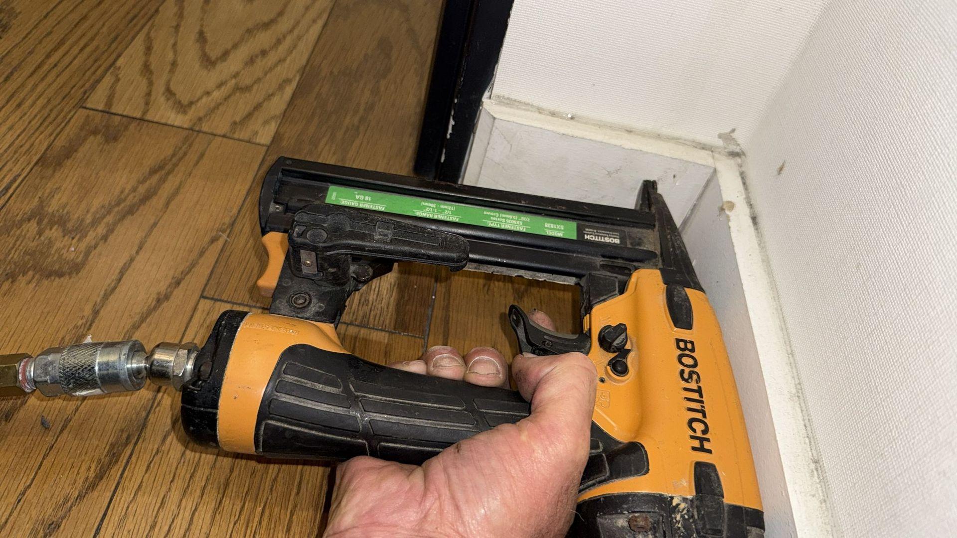 Close-up of a person's hand holding a pneumatic nail gun against wooden floor and white wall.